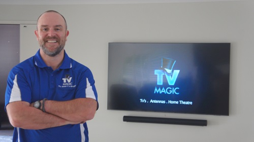 A technician standing in front of a freshly setup and tuned TV mounted on the wall with a sound bar. TV Magic logo on screen