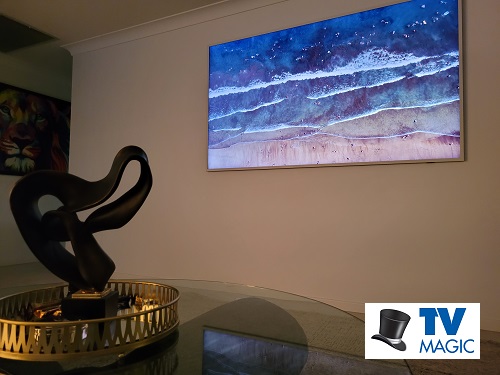 a Samsung Frame TV mounted on a living room wall with a TV Magic logo in bottom right corner