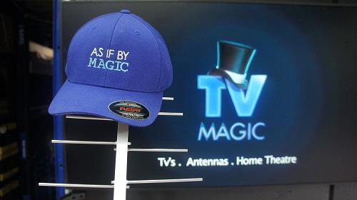 Large TV screen with TV Magic logo on screen. Antenna standing in front of it with a TV Magic cap on top that says "As If By Magic"